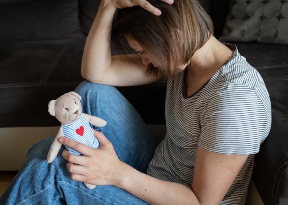 Fejét lehajtó nő a padlón ül, kezében egy szív mintás plüssmacit tart – a baba elvesztésének gyásza